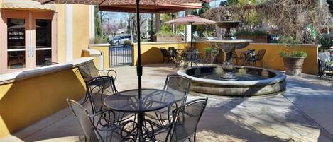 An outdoor patio area with a round table and chairs, a decorative fountain, and a covered seating area with umbrellas.