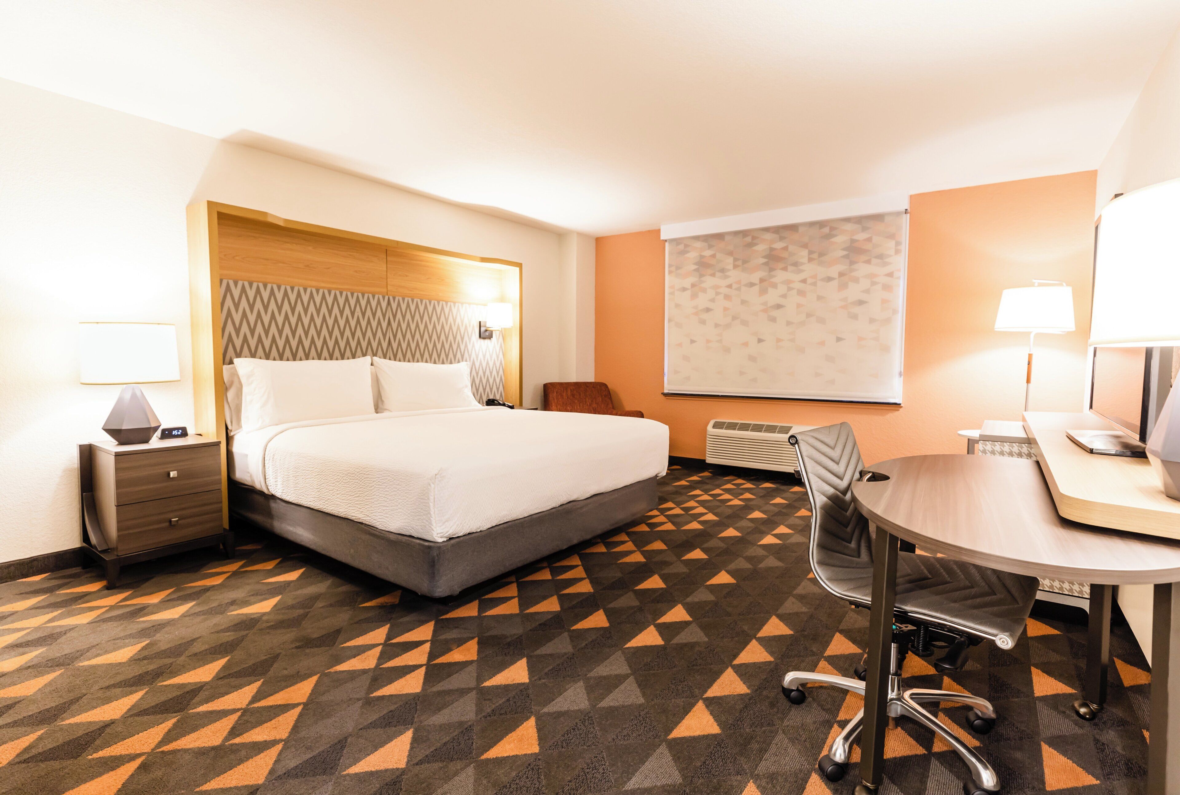Quarto de hotel com cama, mesa de cabeceira, abajur, escrivaninha, cadeira e uma janela grande com persianas.