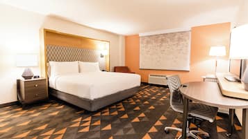 A hotel room with a bed, bedside table, lamp, desk, chair, and a large window with blinds.