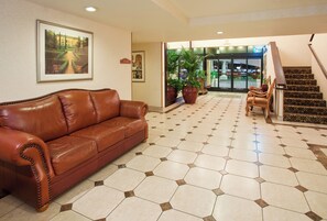 A hotel lobby with a brown leather sofa, a large painting, potted plants, a staircase, and a tiled floor.
