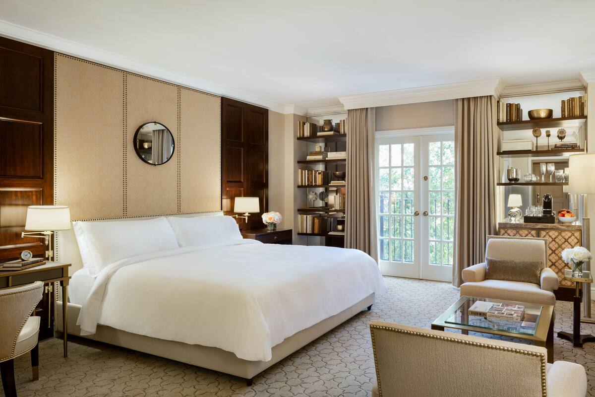 Une chambre d’hôtel spacieuse avec un grand lit, un coin salon avec une table basse, une bibliothèque et une vue à travers les portes vitrées.