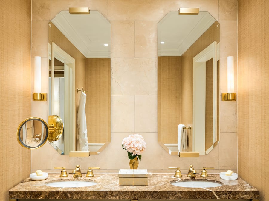 Un baño con dos lavamanos, grifos dorados, un espejo y un florero con flores.