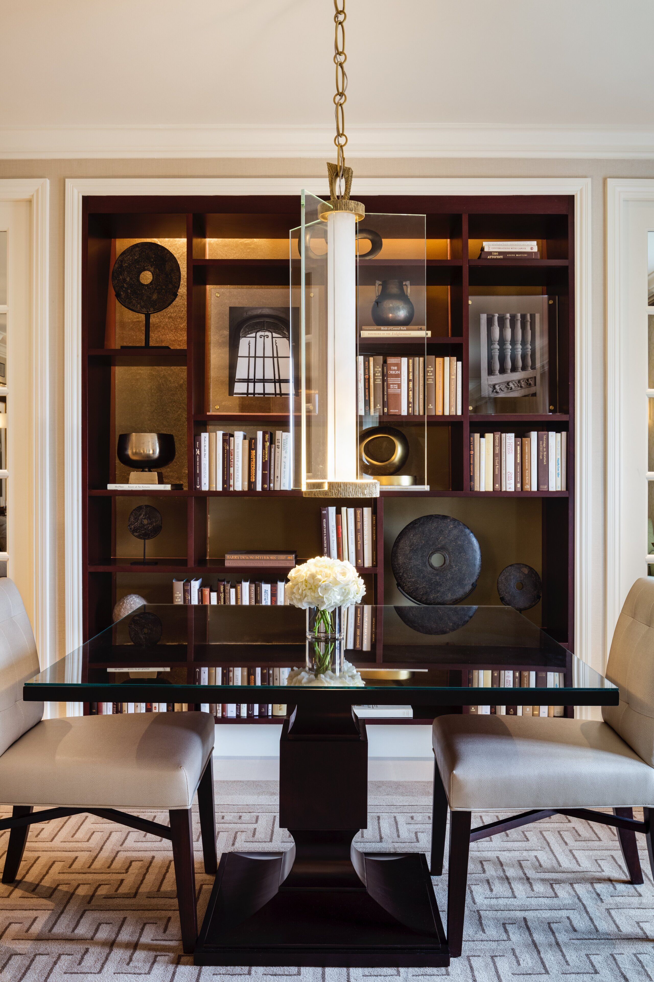 Un comedor moderno con una mesa de cristal, dos sillas, un florero con flores y una estantería llena de libros y objetos decorativos.