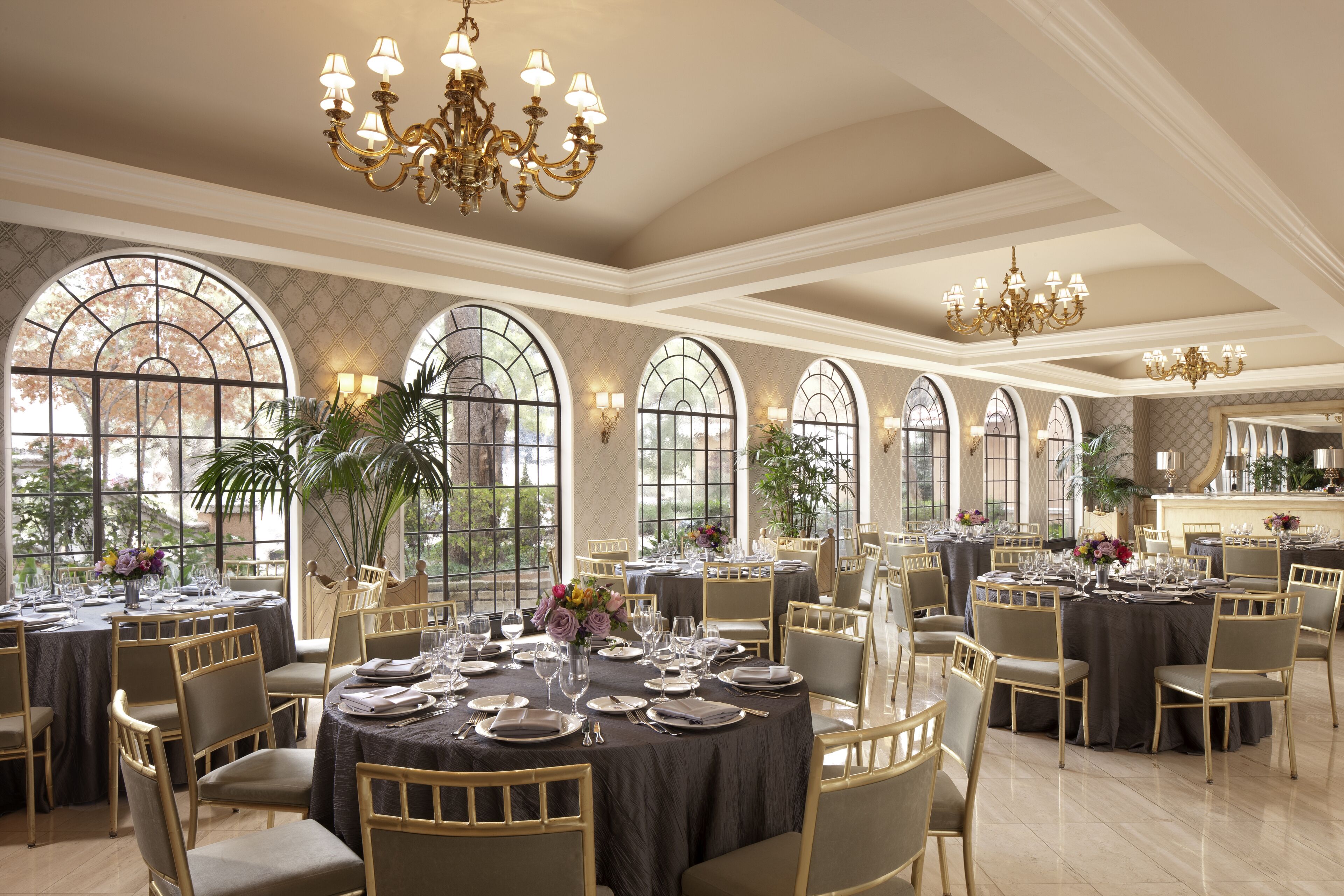 A spacious dining area with multiple round tables set for a meal, large arched windows, and elegant chandeliers.