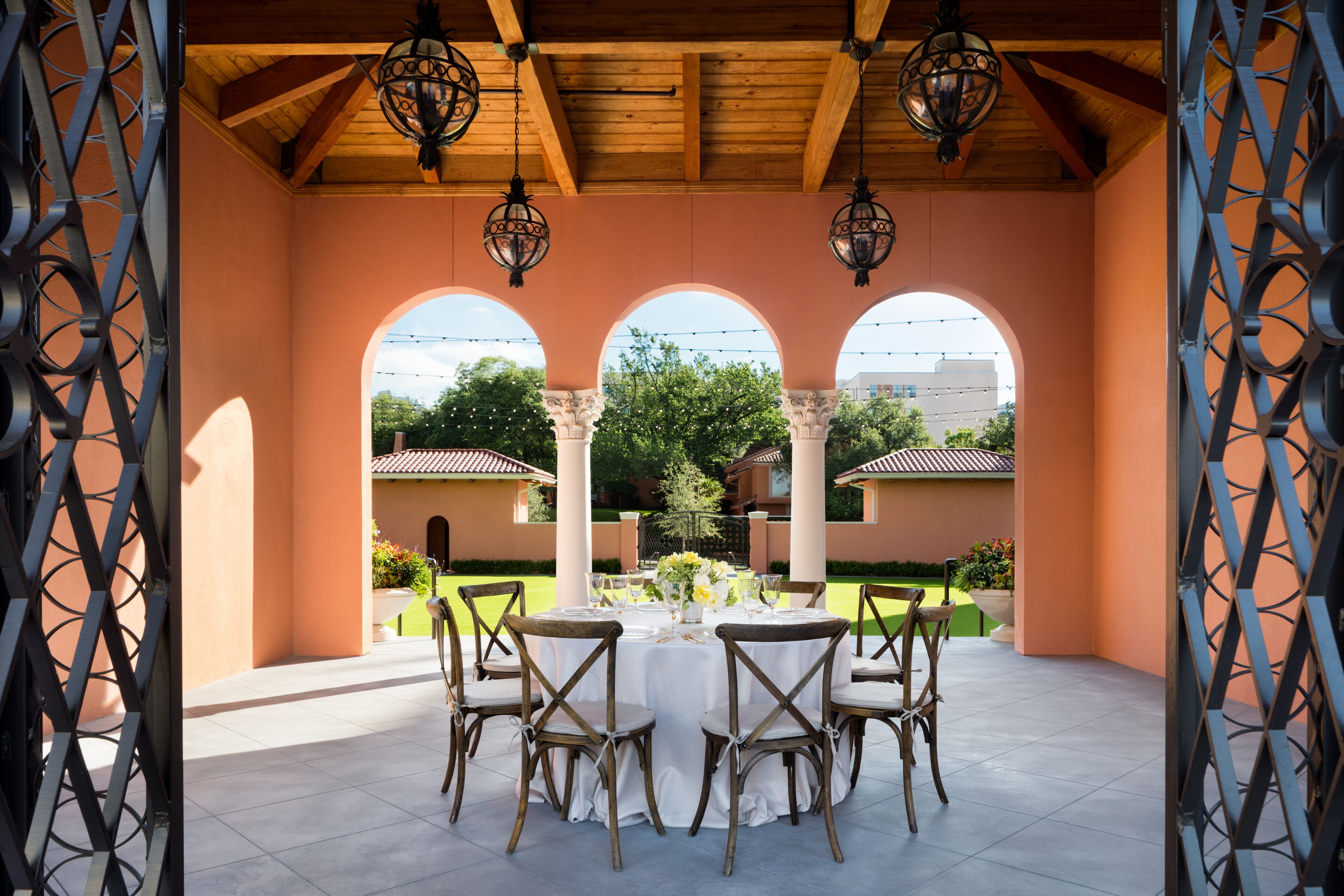 An outdoor patio with a round table set for dining, surrounded by chairs, under a wooden ceiling with hanging lanterns.