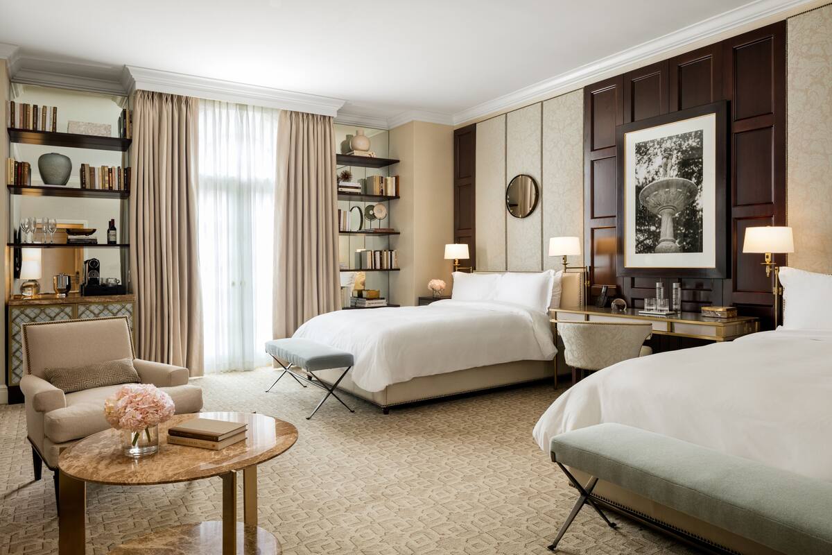 Une chambre d’hôtel avec deux lits, un coin salon comprenant une chaise et une table, une bibliothèque et un grand tableau encadré au mur.