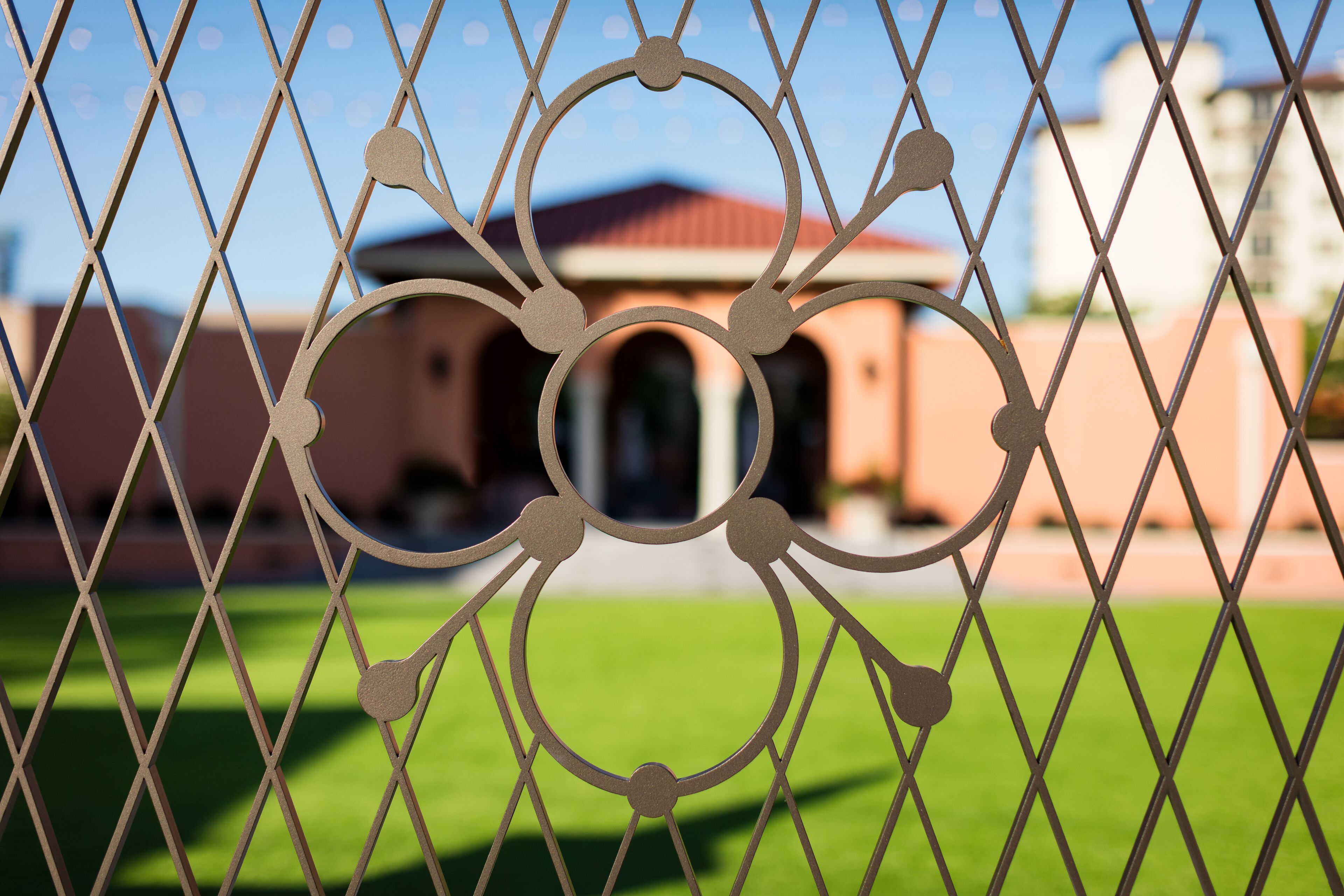 A chain-link fence with a decorative metal design in the foreground, a green lawn, and a building with a red roof in the background.