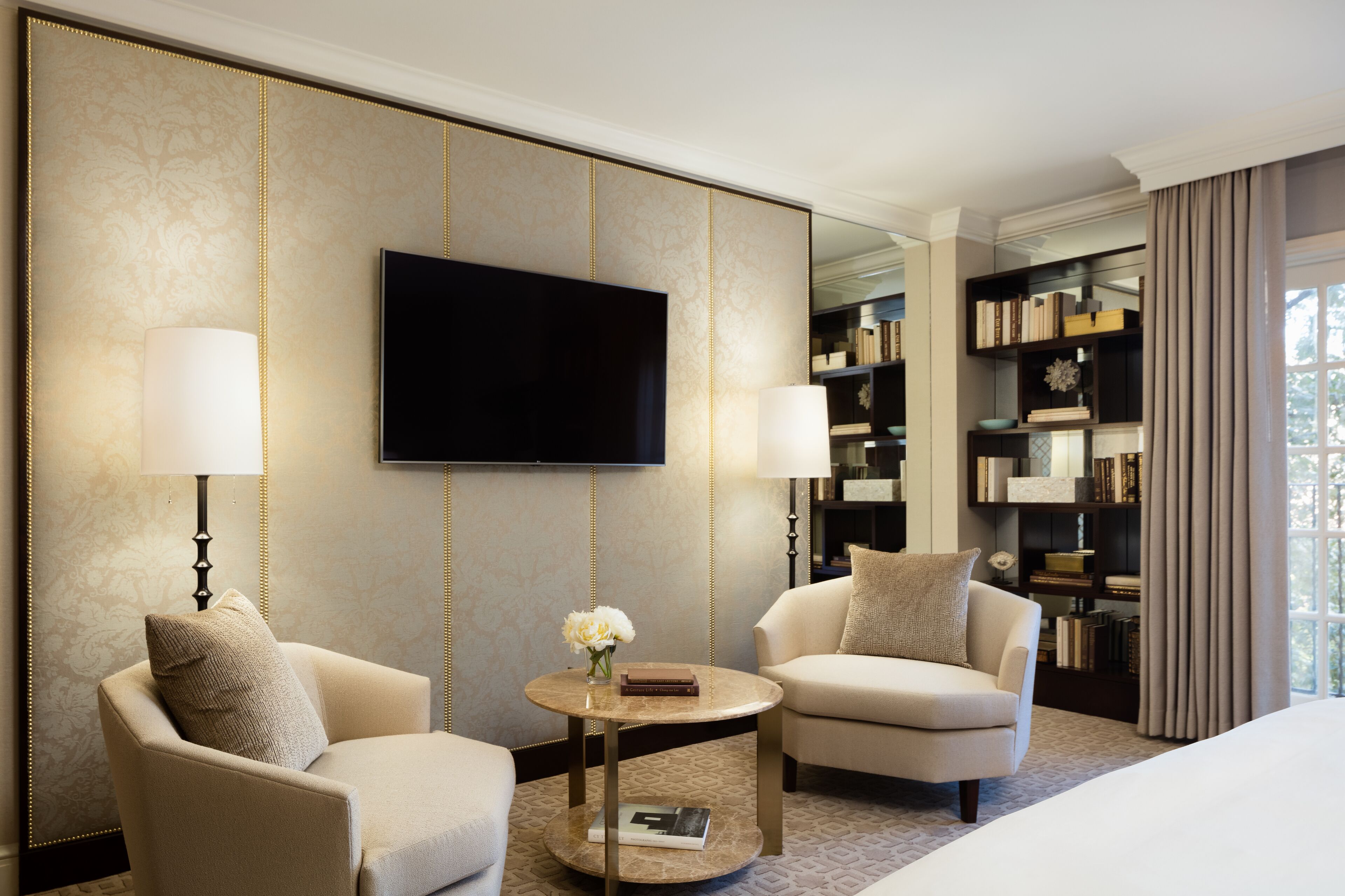A hotel room with two armchairs, a round coffee table, a flat-screen TV, a bookshelf, and a large window with curtains.