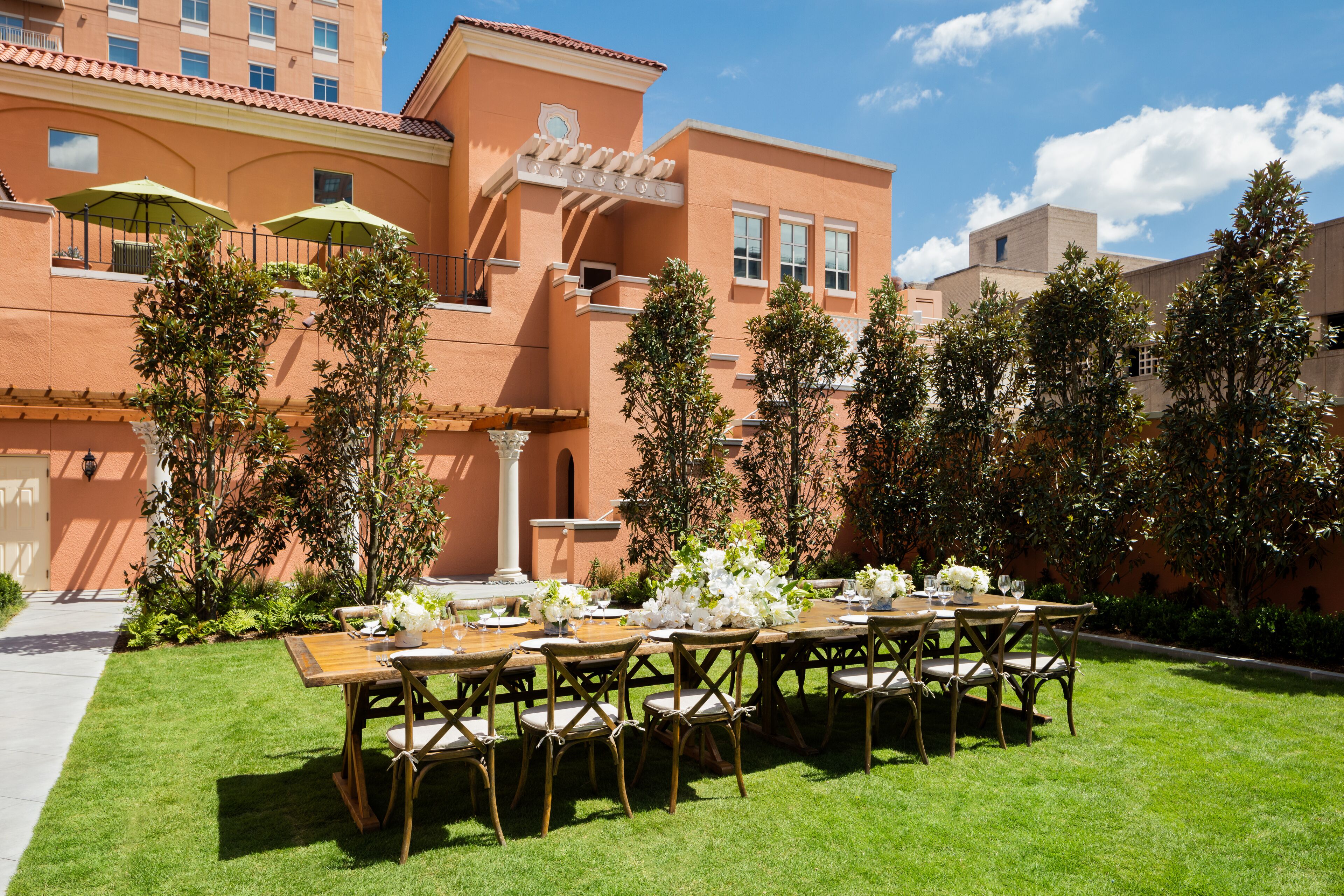 Un espace de restauration en plein air avec une longue table dressée pour un repas, entouré d’une végétation luxuriante et d’un bâtiment à la façade en terre cuite.