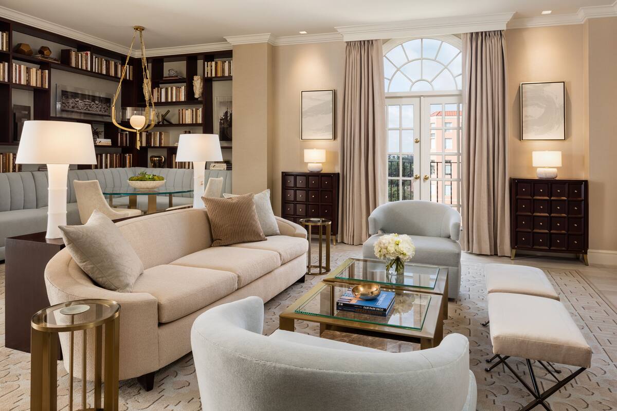 Un salon spacieux avec un canapé beige, une table basse en verre et un fauteuil gris. On y trouve des étagères remplies de livres et un lustre.