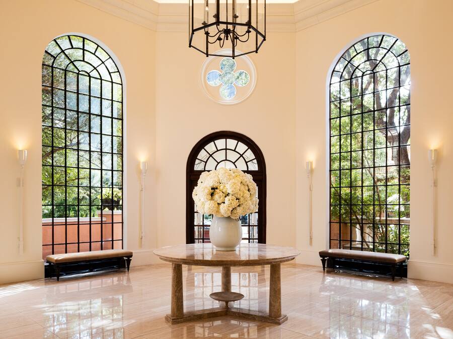 A spacious lobby with a marble floor, a central round table, and three large arched windows with a view of greenery.