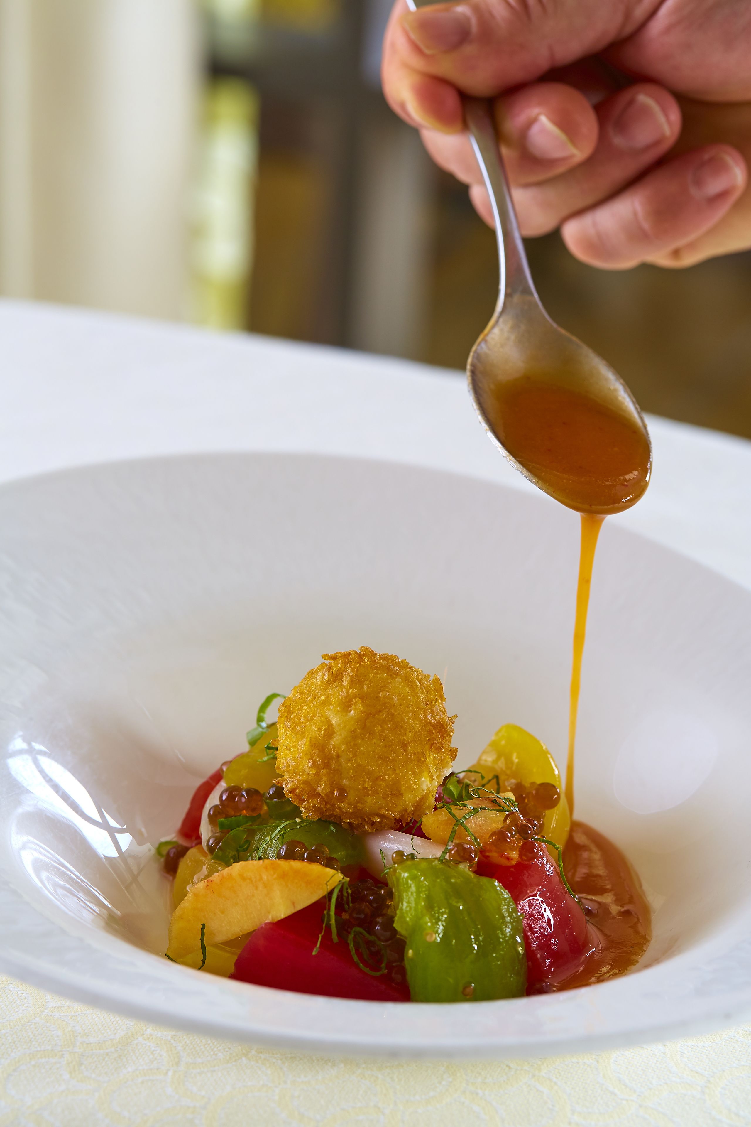 A spoonful of sauce being poured over a dish with various vegetables and a crispy item.