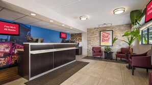 A hotel reception area with a digital screen displaying 'Free Wi-Fi All Rooms,' a reception desk, a 'Red Roof Plus' sign, and a seating area with chairs and a potted plant. - Red Roof Inn PLUS+ Nashville Airport (Nashville)