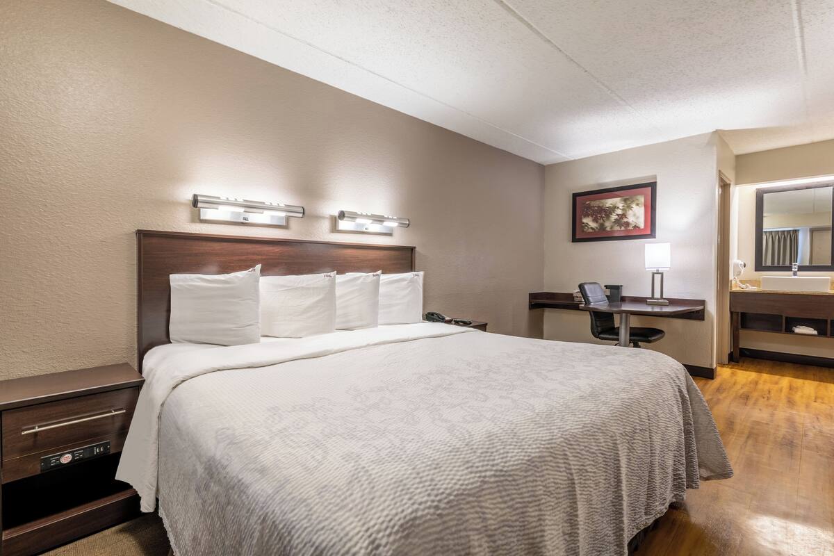 A hotel room with a large bed, a desk, a chair, a mirror, and a wall-mounted light fixture.