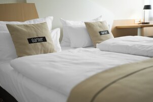 Quarto de hotel com cama bem arrumada, duas almofadas com o nome 'Garner', cabeceira de madeira e mesa de cabeceira pequena com uma luminária.