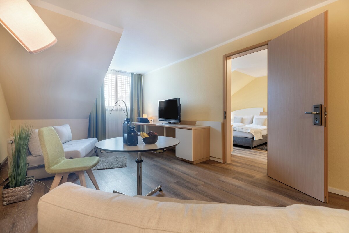 A modern hotel room with a sofa, a small dining table, a TV, and a bed.