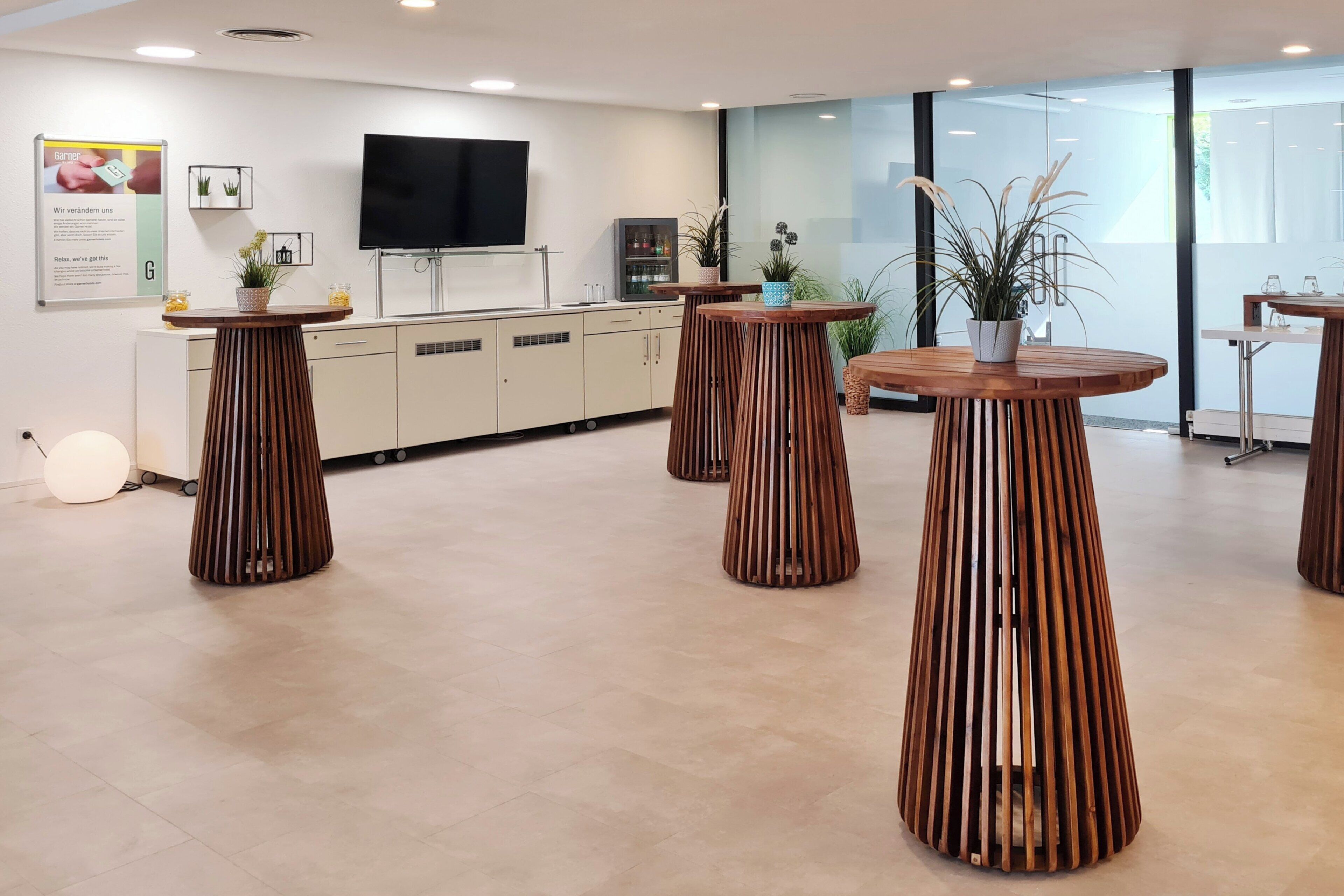 A modern hotel lobby with wooden tables, a flat-screen TV, and potted plants.