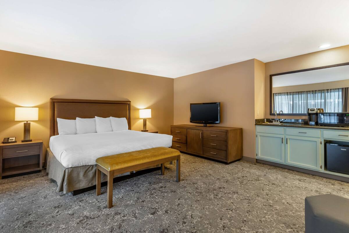 A hotel room with a bed, bedside lamps, a TV, a small table, a bench, a kitchenette with a microwave, and a refrigerator.