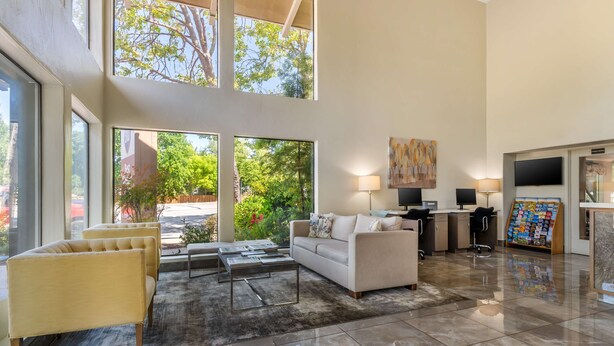 Un espacio habitable amplio con un sofá, sillones, una mesa de centro y un televisor. Hay un escritorio con una computadora y una lámpara. Ventanales grandes ofrecen vistas a árboles y un edificio exterior.