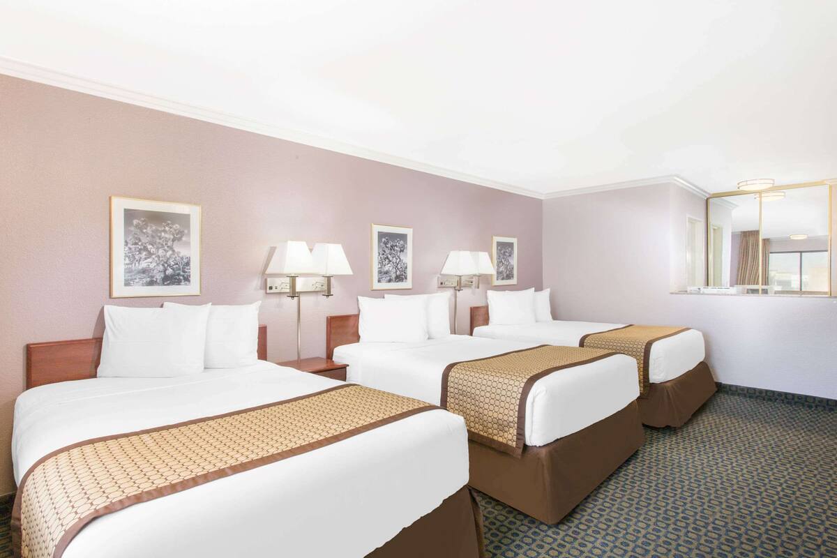 A hotel room with three beds, each with a brown bedspread, white pillows, and bedside lamps. There are framed pictures on the wall and a large mirror on the right side.