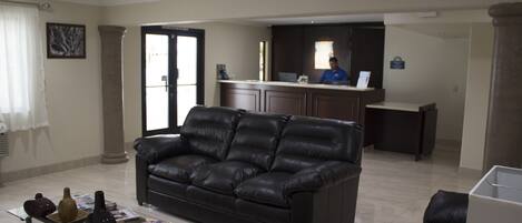 Uma sala de estar com um sofá de couro preto, uma mesa de centro de vidro com objetos decorativos e um balcão de recepção com uma pessoa atrás.