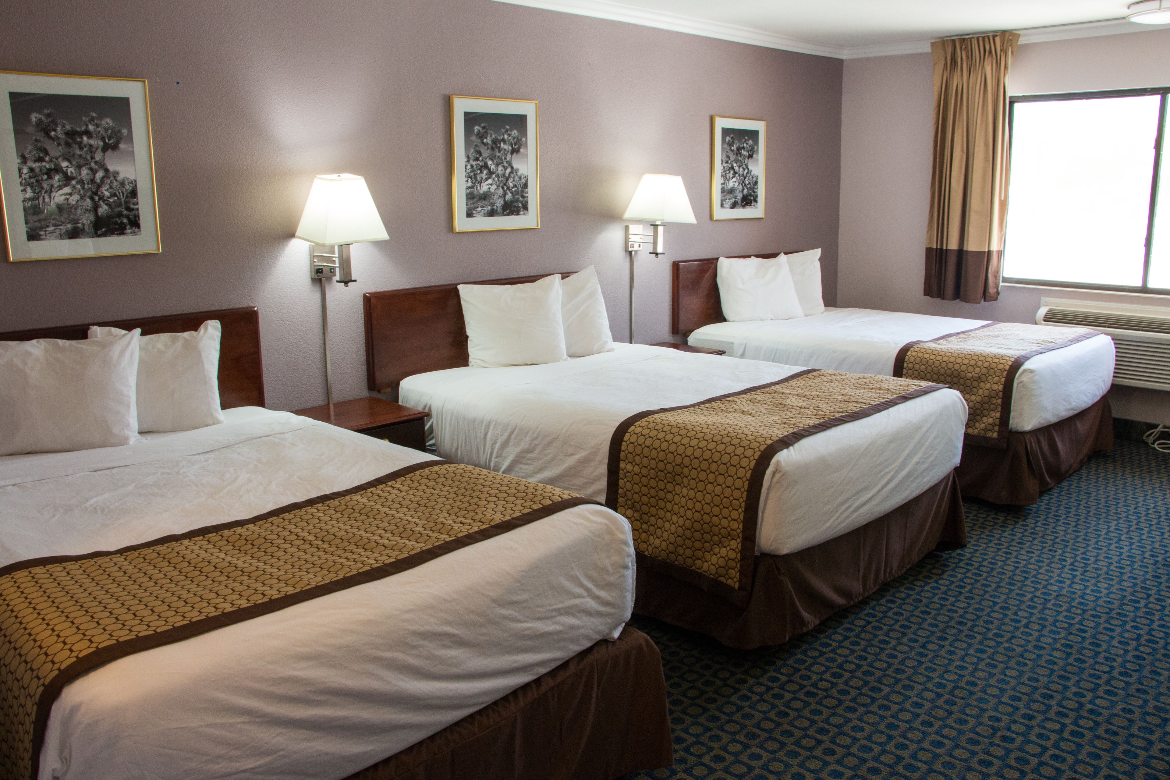 Une chambre d’hôtel avec trois lits, chacun doté d’une tête de lit marron et de draps blancs. On y trouve deux lampes fixées au mur et des tableaux encadrés accrochés au mur.