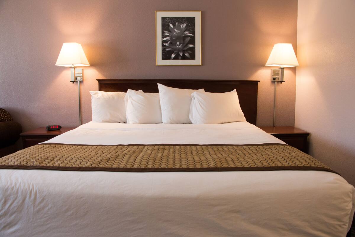 A hotel room with a neatly made bed, two bedside lamps, a framed artwork on the wall, and a digital clock on the nightstand.