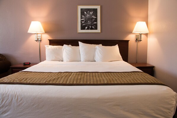 A hotel room with a neatly made bed, two bedside lamps, a framed artwork on the wall, and a digital clock on the nightstand.