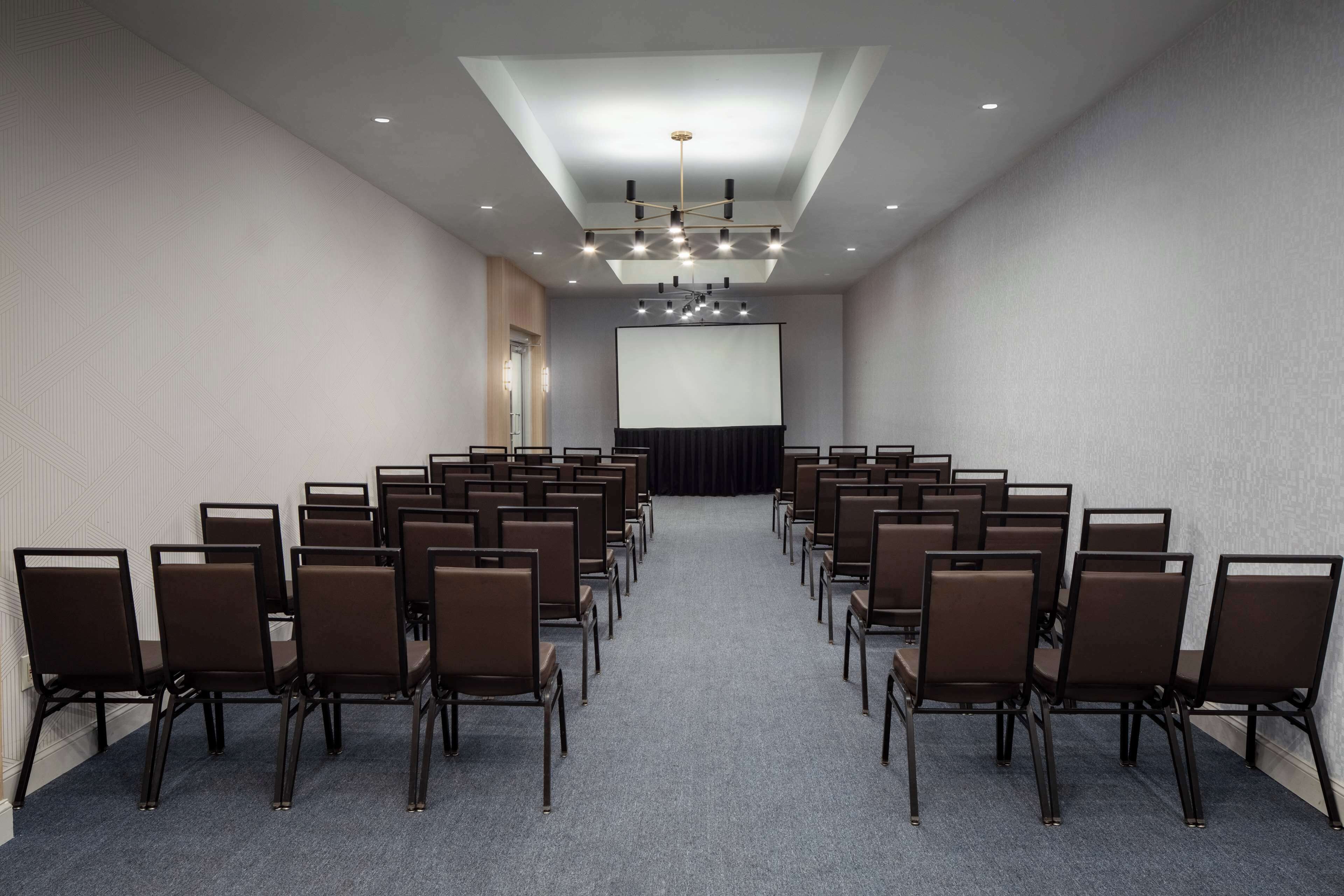 A conference room with rows of chairs facing a large screen, a ceiling light, and a door on the left.