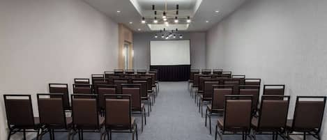 A conference room with rows of chairs facing a large screen, a ceiling light, and a door on the left.
