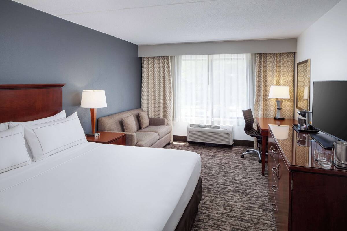 A hotel room with a large bed, a sofa, a desk with a television, and a window with curtains.