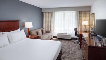 A hotel room with a large bed, a sofa, a desk with a television, and a window with curtains.