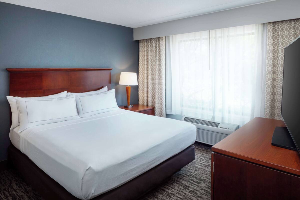 A hotel room with a large bed, a nightstand with a lamp, a TV, and a window with curtains.