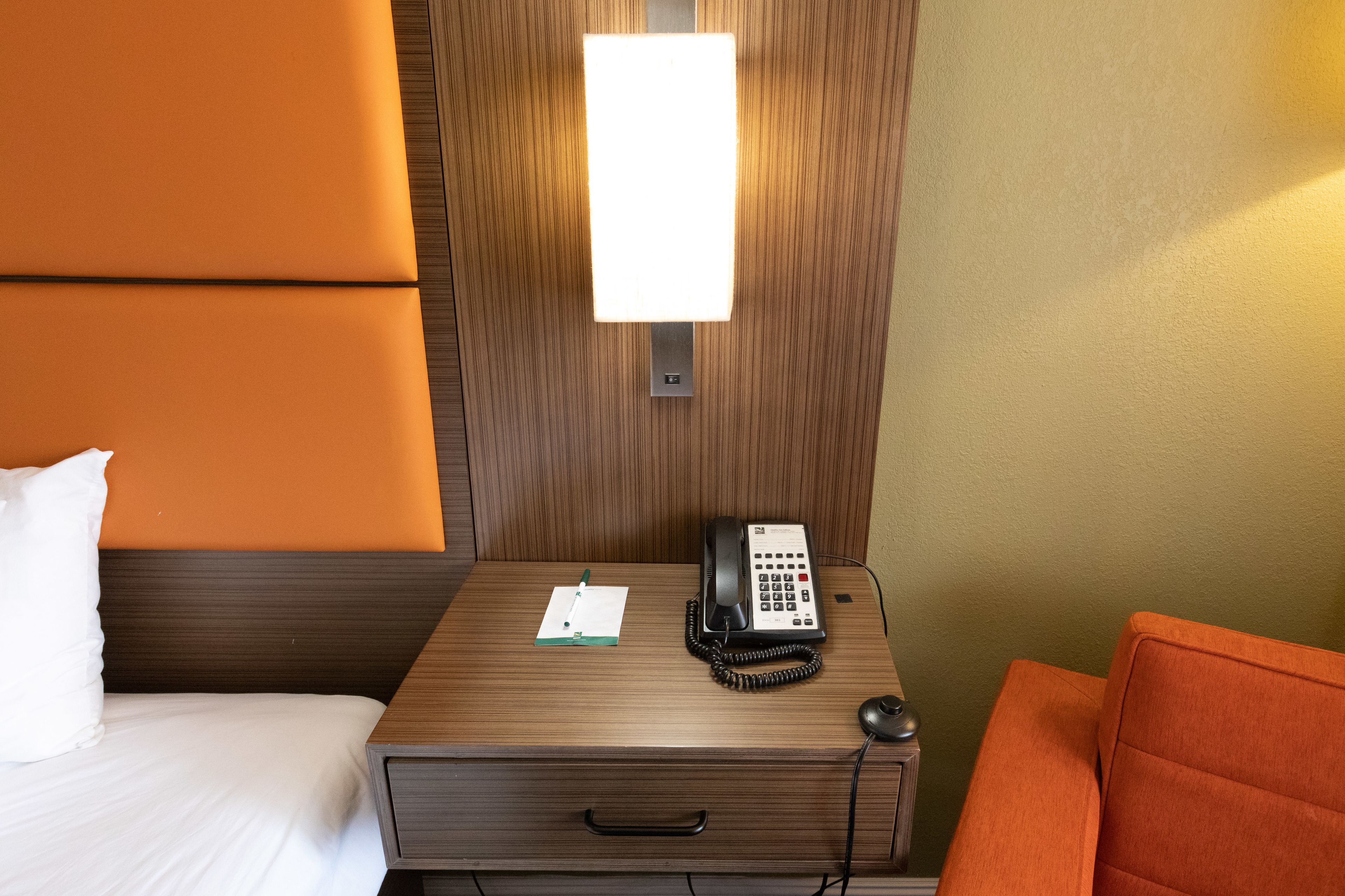 Una habitación de hotel con una mesita de noche de madera, un teléfono, una libreta y una silla.