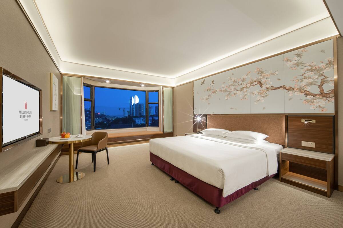 Ein modernes Hotelzimmer mit einem großen Bett, einem Schreibtisch und Blick auf die Stadt bei Abenddämmerung.