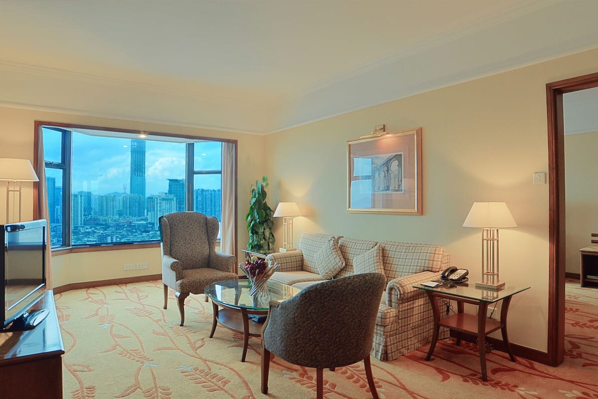 Ein Hotelzimmer mit großem Fenster und Blick auf die Stadt, ausgestattet mit einer Couch, Sesseln, einem Glastisch, einem Schreibtisch mit Lampe und einem Telefon.