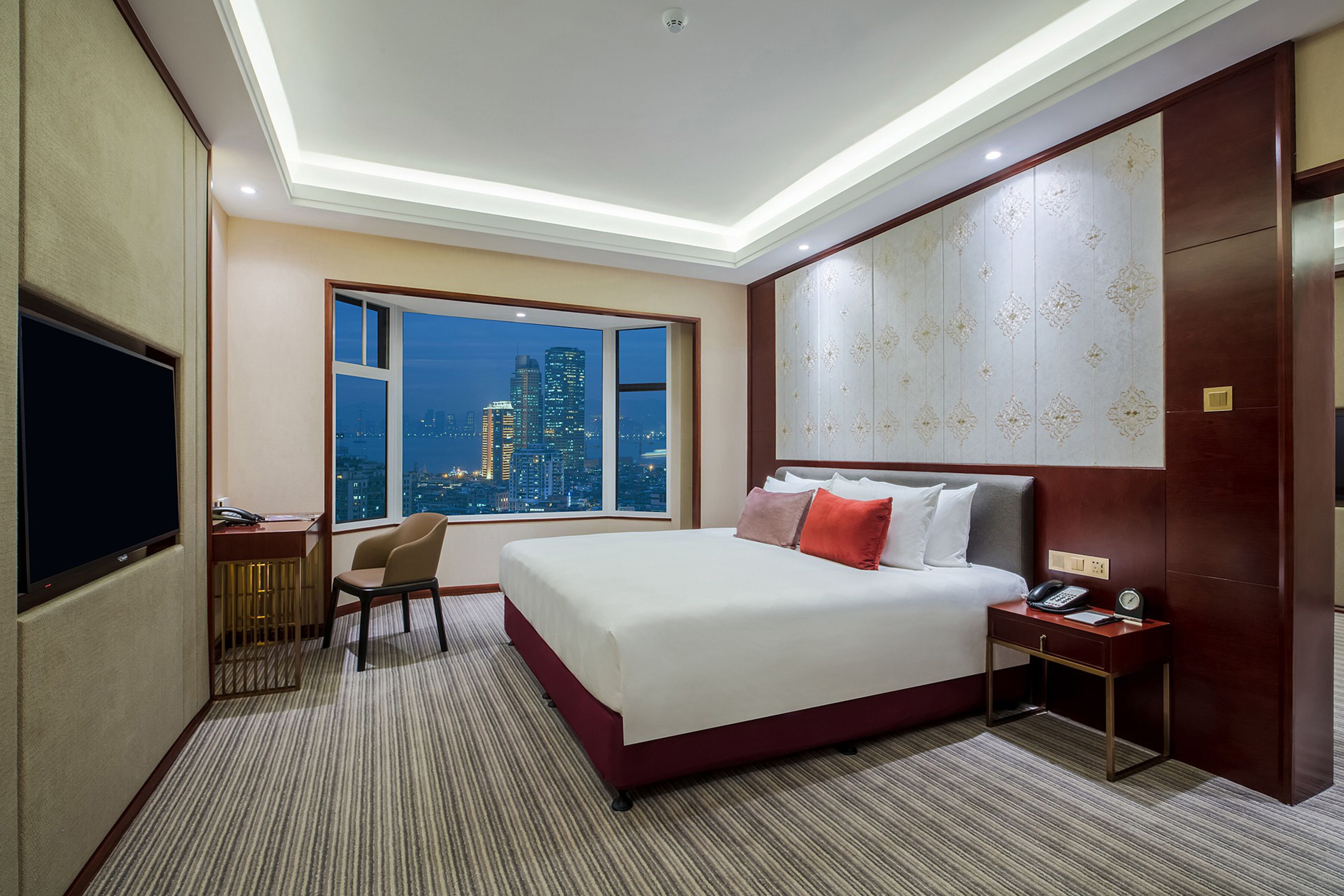 A hotel room with a large bed, a desk with a chair, a television, and a view of a cityscape at night.