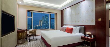 A hotel room with a large bed, a desk with a chair, a television, and a view of a cityscape at night.