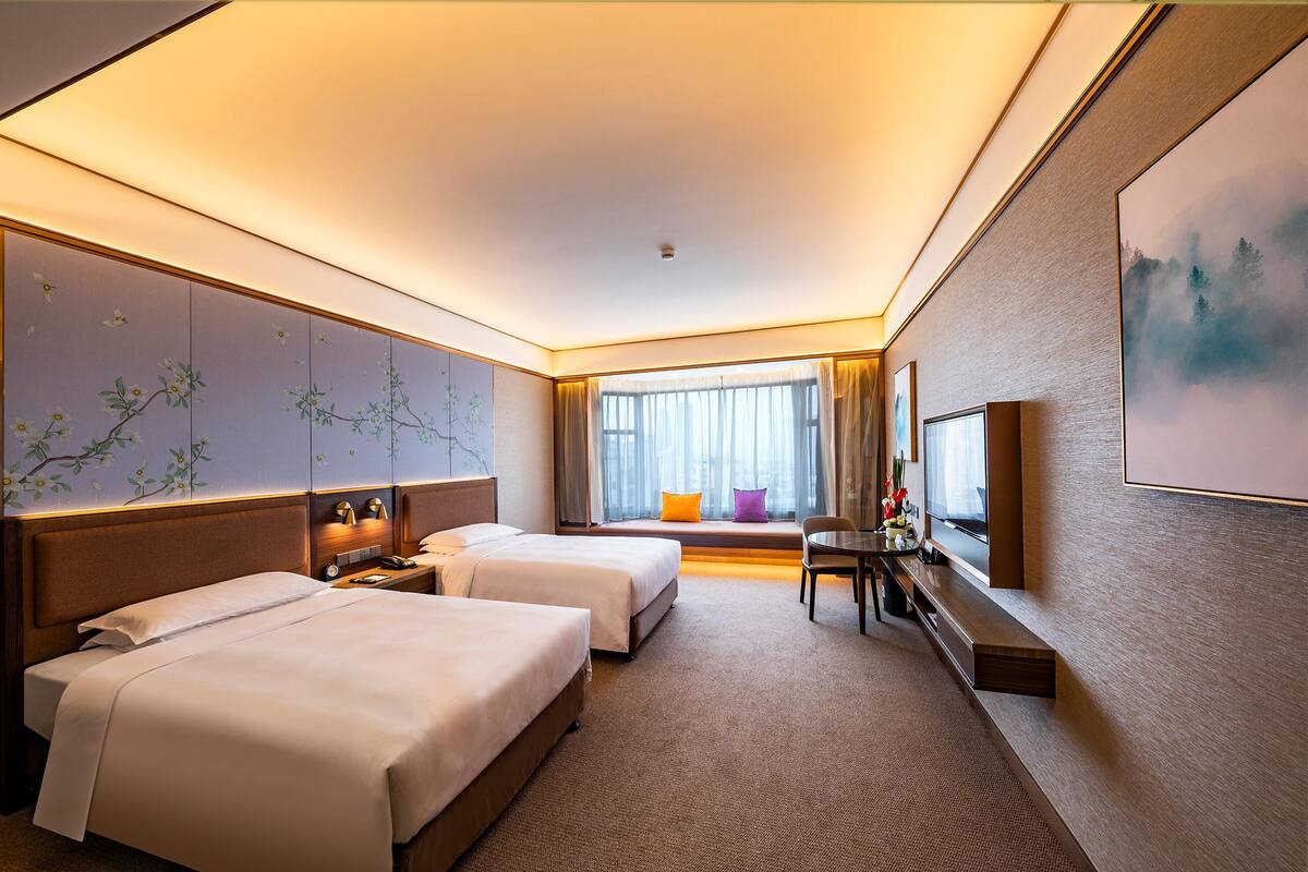 Ein Hotelzimmer mit zwei Betten, einem Schreibtisch, einem Stuhl, einem Fernseher und einem großen Fenster mit Vorhängen.