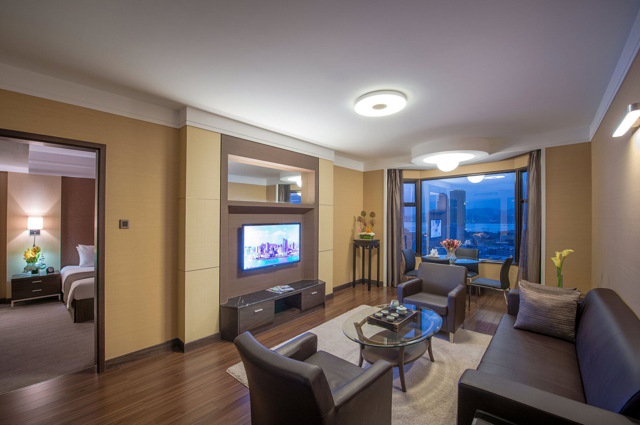 A modern hotel room with a flat-screen TV, comfortable seating, a small table, and a view of the city.