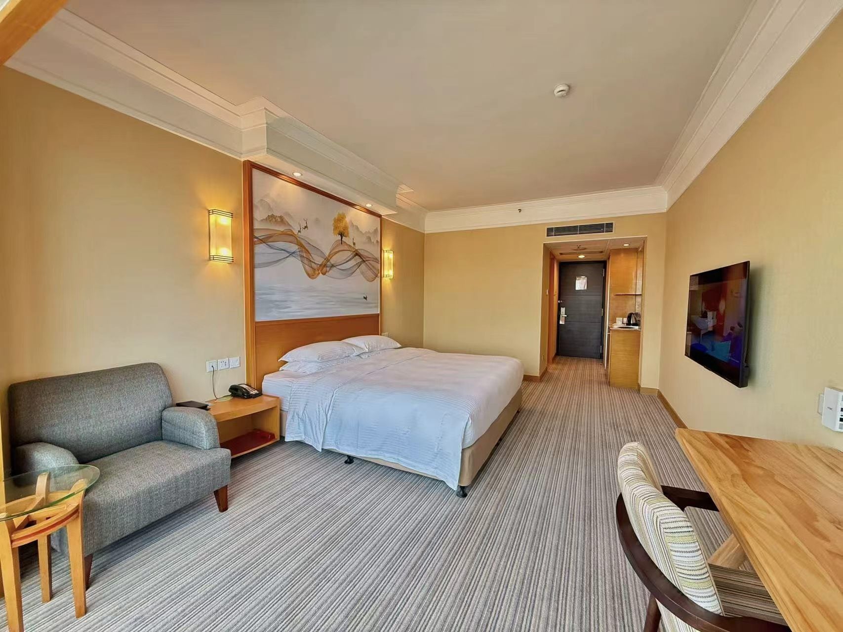 A hotel room with a bed, a sofa, a desk, a painting on the wall, and a door leading to another room.