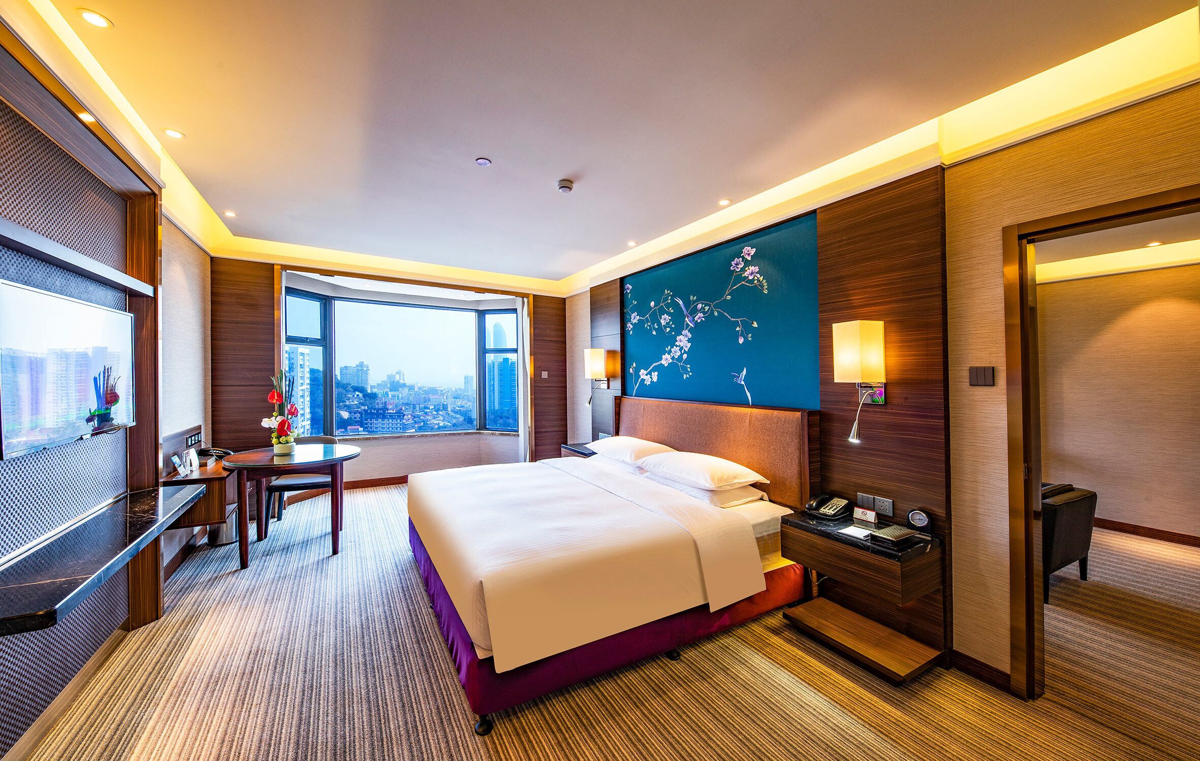 A hotel room with a large bed, a desk with a lamp, a TV, and a city view.