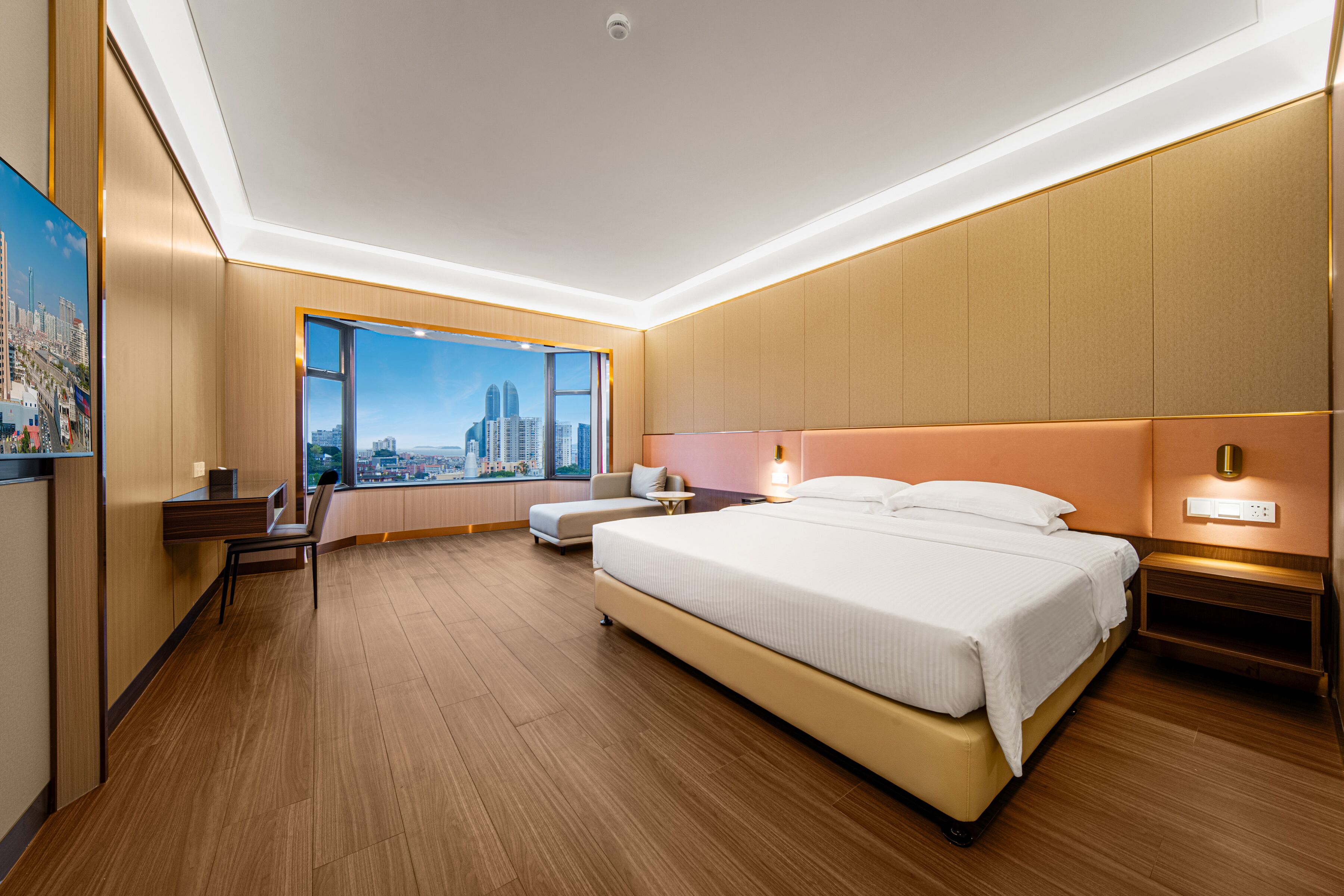 A modern hotel room with a large bed, a desk, and a view of a cityscape through a large window.