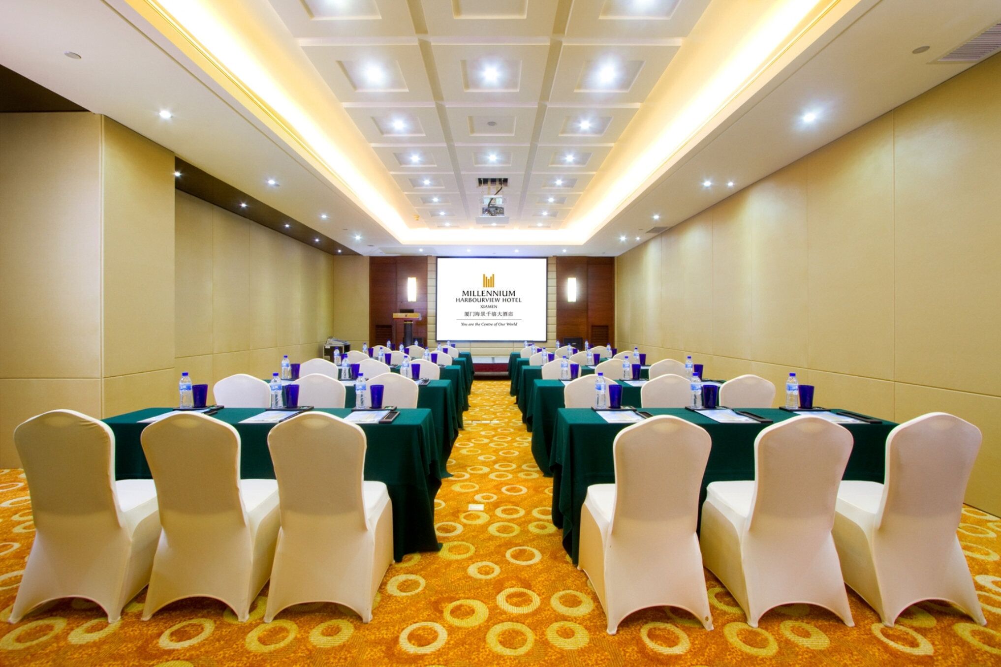 A conference room with rows of white chairs, green tables, and a carpeted floor. A screen displays the name of the hotel.