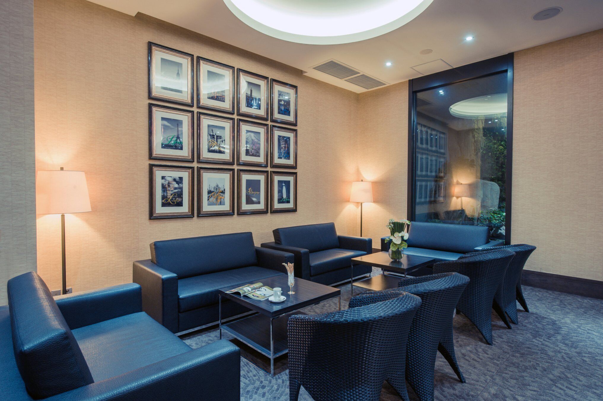 A modern lounge area with sofas, a coffee table, and wicker chairs. There are framed pictures on the wall and a large window providing a view.
