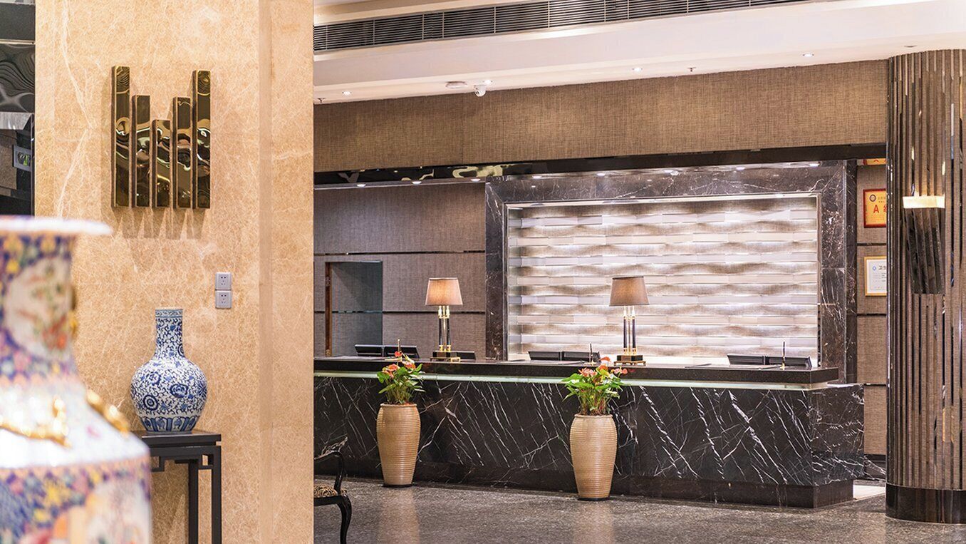 A reception area with a marble reception desk, two potted plants, a wall-mounted mirror, and a patterned wall.