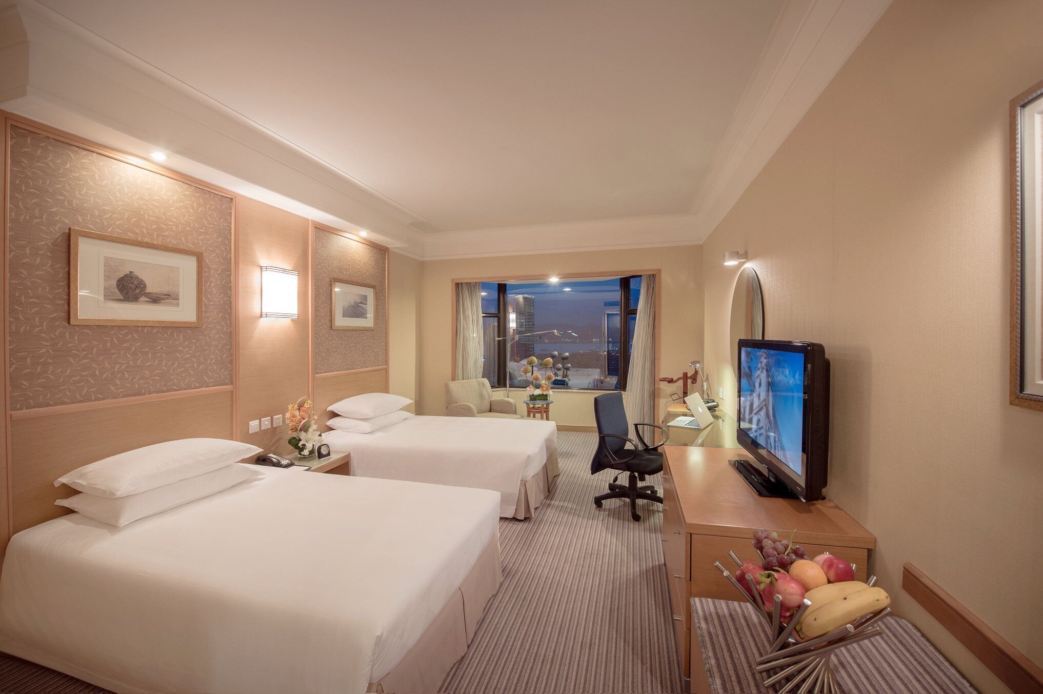 Habitación de hotel con dos camas, un escritorio con televisor, una silla y una ventana con vista a la ciudad.