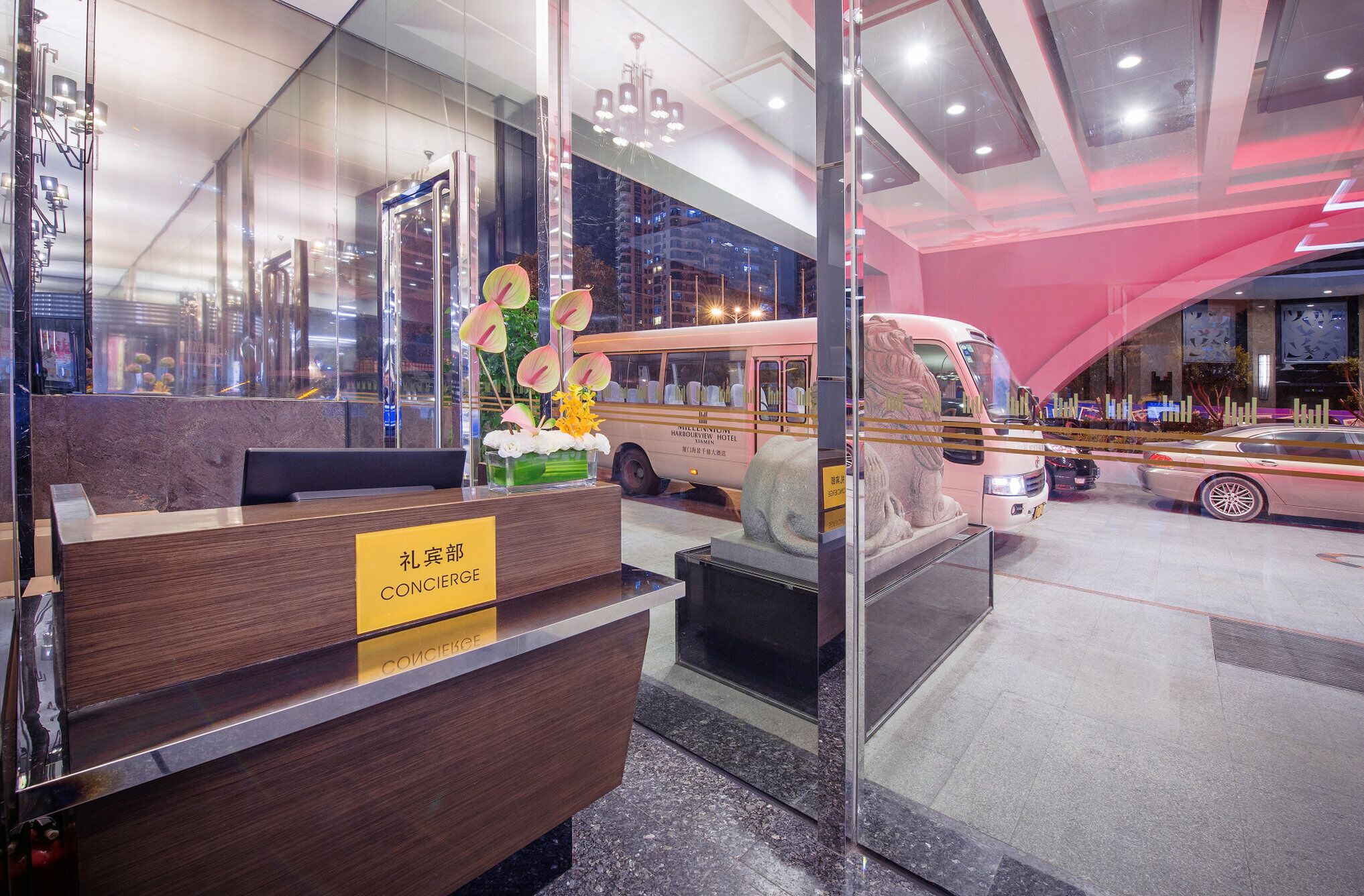 A hotel reception area with a wooden reception desk, a glass partition, and a view of a parked vehicle outside.