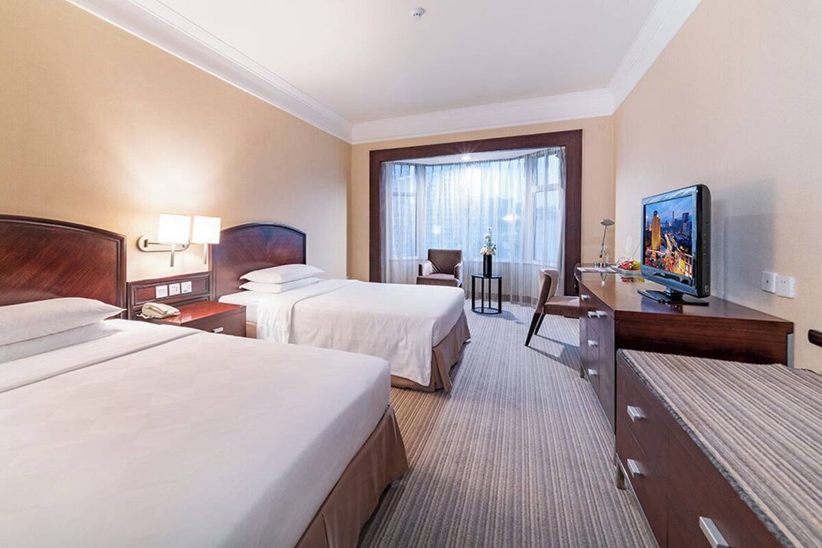 Ein Hotelzimmer mit zwei Betten, einem Schreibtisch mit Fernseher, einem Sessel und einem großen Fenster mit Vorhängen.