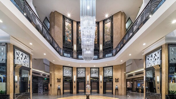 Een ruime lobby met een centrale kroonluchter, meerdere liften en een marmeren vloer.